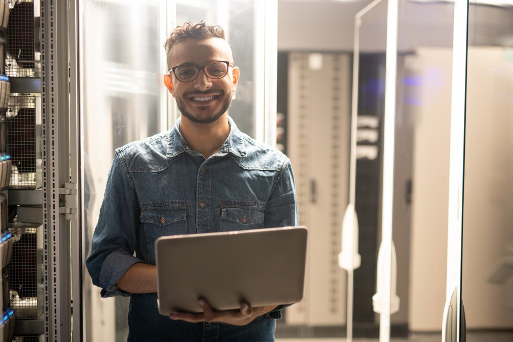 T2G - Portrait of cheerful satisfied young middle-eastern server engineer in glasses standing in datacenter room and using laptop
